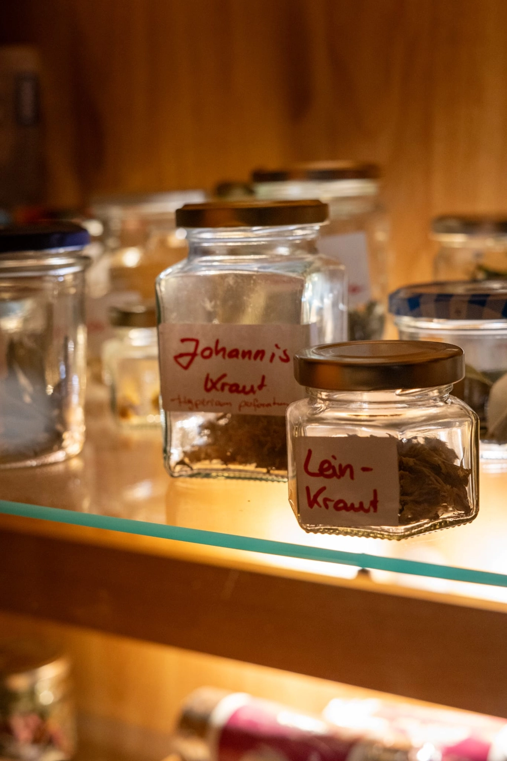 Energie Stüberl - Marianne Roßmadl - Energetische Heilarbeit und Massage - Gläser mit handgeschriebenen Etiketten mit der Aufschrift „Johannis Kraut“ und „Lein-Kraut“ auf einem Glasregal mit warmem Holzhintergrund