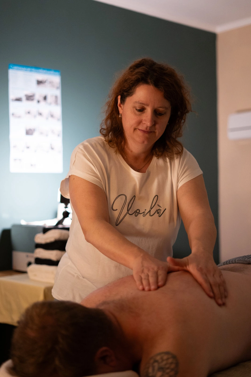 Energie Stüberl - Marianne Roßmadl - Energetische Heilarbeit und Massage - Frau im weißen „Voilà“-T-Shirt gibt einem Mann, der mit dem Gesicht nach unten auf einer Massageliege liegt, eine Rückenmassage