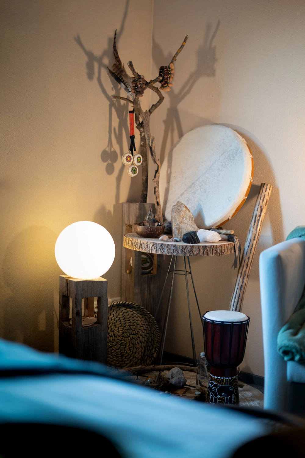 Energie Stüberl – Marianne Roßmadl – Energetische Heilarbeit und Massage – Gemütliche Ecke mit einer runden, leuchtenden Lampe auf einem Holzständer, einem kleinen Holztisch mit Steinen und Kristallen, einer großen Trommel, einem Hebelstab und einem geflochtenen Korb vor einer beigen Wand