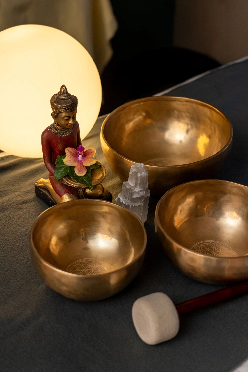 Energie Stüberl – Marianne Roßmadl – Energetische Heilarbeit und Massage – Drei goldene Klangschalen mit einem Holzschlägel, eine Statue mit einer Blume und ein Kristall auf dunklem Stoff neben einem leuchtenden runden Licht
