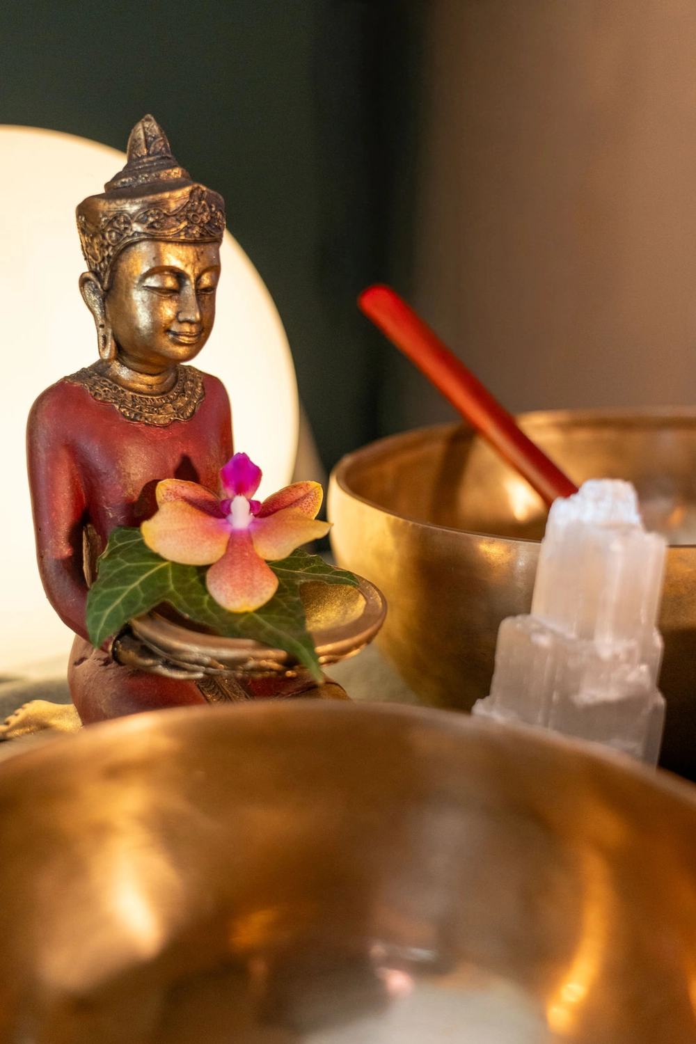 Energie Stüberl - Marianne Roßmadl - Energetische Heilarbeit und Massage - Dekorative Buddha-Statue mit einem Teller, der eine rosa-orangefarbene Orchideenblüte und grüne Blätter trägt, umgeben von goldenen Schalen und einem weißen Kristall