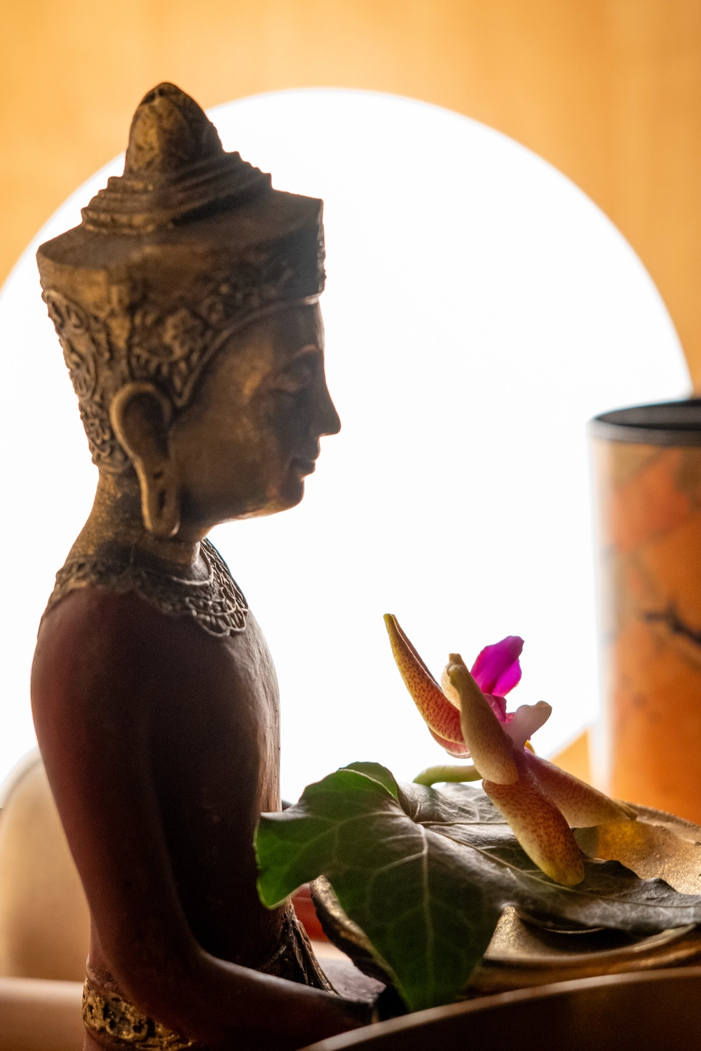 Energie Stüberl - Marianne Roßmadl - Energetische Heilarbeit und Massage - Seitenansicht einer dekorativen Buddha-Statue, die ein Blatt mit einer kleinen Blüte hält, vor einem leuchtenden kreisförmigen hellen Hintergrund